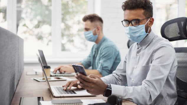 Two young business men, one white and one multi-racial, working at computers while wearing face masks.