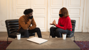 Two colleagues, a man and woman, both wear masks and discuss something over coffee