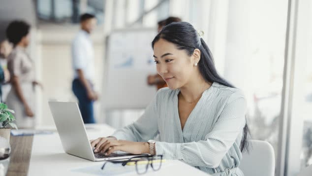 Asian woman working on laptop. Businesswoman busy working on laptop computer at office with colleagues in the background.