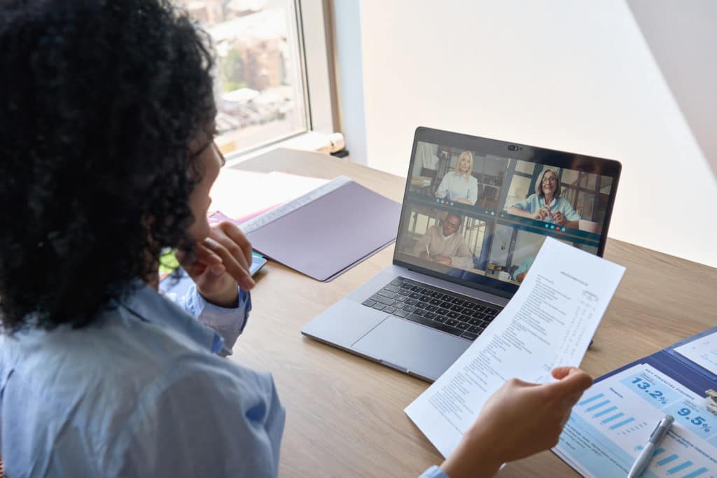 African American businesswoman ceo holding papers having videocall on yearly financial report with multiethnic colleagues using laptop in modern corporation office. Over shoulder view.