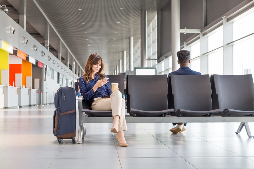 Young woman waiting for flight at the airport lounge. Businesswoman sitting on a bench with coffee and using a mobile phone.
