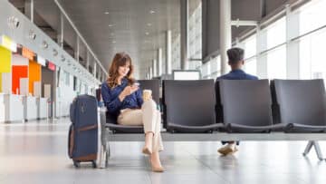 Young woman waiting for flight at the airport lounge. Businesswoman sitting on a bench with coffee and using a mobile phone.